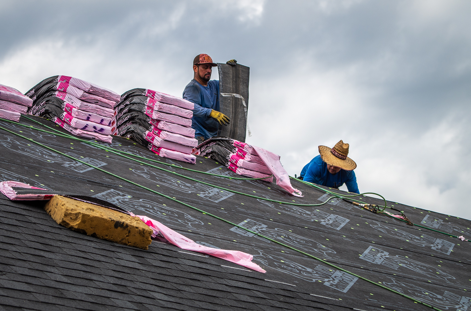 Roofing contractors install shingles on a sloped roof, with pink shingle bundles nearby, under a cloudy sky.
